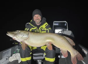 Winter night pike fishing – angler holding a massive pike on a boat in extreme cold